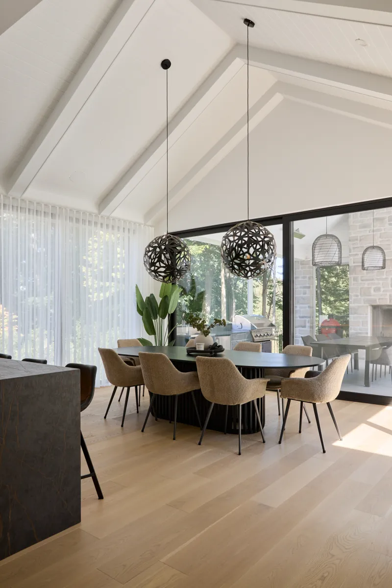 Salle à manger vitrée avec table ovale, luminaire suspendu et plafond cathédrale à poutres blanches.